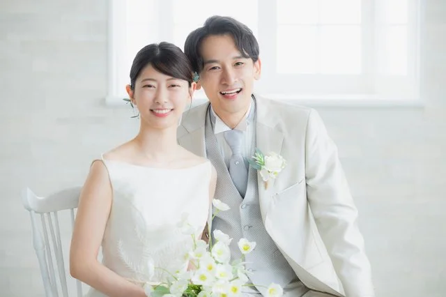 白い壁と窓を背景に、花束を持って寄り添うウェディング姿の男女